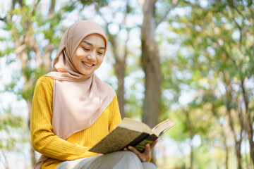 Wall Mural - Happy cheerful Asian beautiful Muslim woman reading a book - Koran under the tree. Female muslim student studying an Islam Koran.