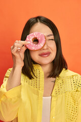 portrait of young, pretty, asian girl in yellow blouse, holding pink donut, posing against orange st