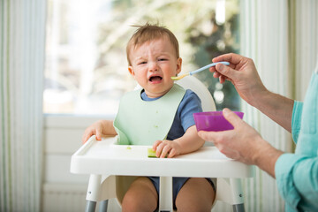 father feeding his cute baby with first solid food, infant sitting in high chair. child doesn't want