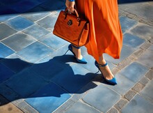 Women's Legs In Orange Shoes. Bright Orange Shoes And Handbag. Stylish Slim Girl In Soft Blue Warm Coat, Gray Warm Dress And Heels Walking On The Rainy Wet Street.