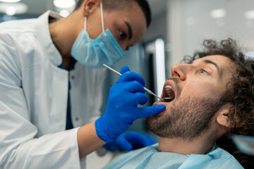 Young man with mouth open having dental exam during appointment at dentist's office, getting his teeth checked by female dentist with surgical mask and gloves at dental clinic.