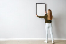 Young Woman Hanging Blank Frame On Light Wall At Home, Back View