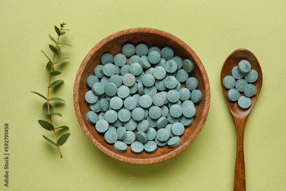 Bowl with pills, spoon and plant branch on green background