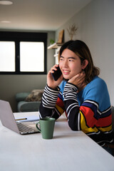 Young taiwanese man on a phone call working at home with a laptop and a cup of coffee.