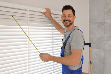 Canvas Print - Worker in uniform using measuring tape while installing horizontal window blinds indoors