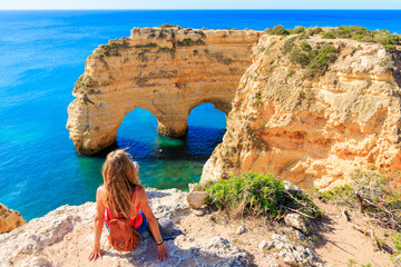 Wall Mural - Woman tourist looking at heart shape rock formation,  coast of the Algarve- Tourism,  travel,  vacation in Europe (natural cave or arch on praia da marinha)- Portugal