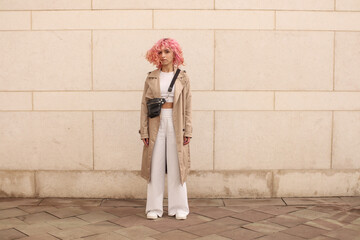 Wall Mural - Portrait shooting of a stylish girl with pink hair. Beige shades. Trends of spring and summer 2023.Beige Trench cloak with white top and pants. Black small belt bag