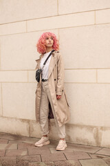 Wall Mural - Portrait shooting of a stylish girl with pink hair. Beige shades. Trends of spring and summer 2023. Beige trench cloak with white t-shirt and beige pants. Black small belt bag