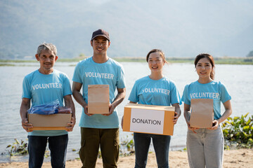 Portrait Team of volunteers wearing volunteer t-shirt,  holding donations box, donations bag and looking at camera, standing on river background