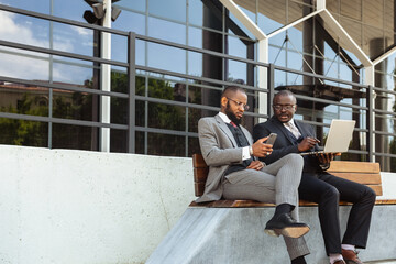 Sticker - Business meeting of friends outdoors. Two dark-skinned men in suits are sitting on a bench near a city building with a laptop and talking. Partnership and corporate relations