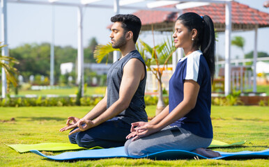 Side view shot of Indian young couple doing yoga or meditation at park during morning - concept of mindfulness, training and tranquility