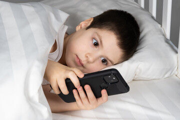 Wall Mural - A 5 year old boy plays games on his phone while lying in bed. Gadgets in bed before bedtime in a child