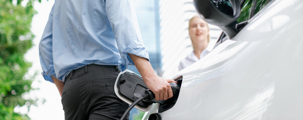 Closeup focus hand insert EV charger to electric vehicle at public charging point in car park with blur business people in backdrop, eco-friendly lifestyle by rechargeable car for progressive concept.