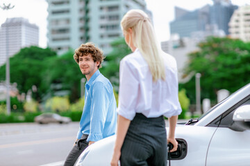 Progressive businessman and businesswoman with electric car parking and connected to public charging station before driving around city center. Eco friendly rechargeable car powered by clean energy.