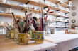 Lots of brushes resting on a table and some pottery behind. Art classes. Instruments and materials in the studio