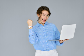 Young happy excited business woman executive holding laptop computer raising fist in yes gesture celebrating online win, business success, good result or reward standing isolated on grey background.