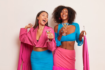 Two ladies having fun inside studio, both girls hold glass of cocktail in one hand and pink dress on wire hangers in another, happy time concept, copy space, high quality photo