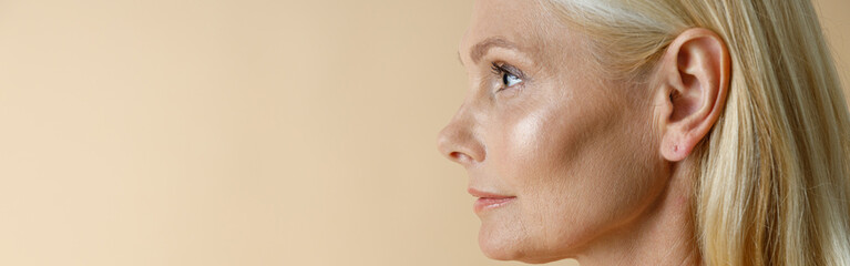 Wall Mural - Side view portrait of pretty mature blonde woman in white underwear looking away, standing isolated over beige background