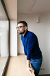 © Cavan Images - brunette man in glasses and a blue sweater looks out the window alone