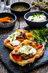 Wall Mural - Tasty sandwiches - toasted bread with burrata cheese, anchovies, bell pepper, olives, tomatoes and thyme on wooden table
