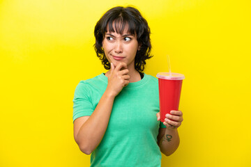 Wall Mural - Young Argentinian woman holding a soda isolated on yellow background having doubts and thinking