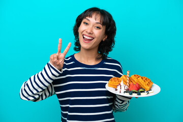 Wall Mural - Young Argentinian woman holding waffles isolated on blue background smiling and showing victory sign