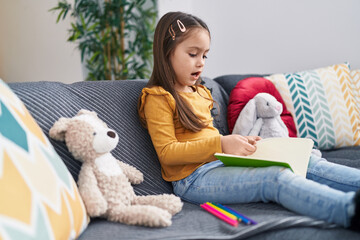 Wall Mural - Adorable hispanic girl drawing on notebook sitting on sofa at home