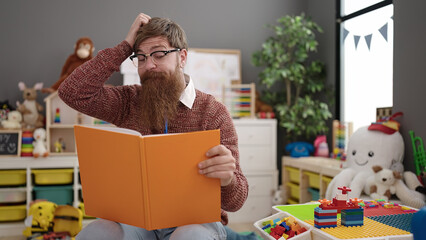 Canvas Print - Young redhead man preschool teacher reading book sitting on chair at kindergarten