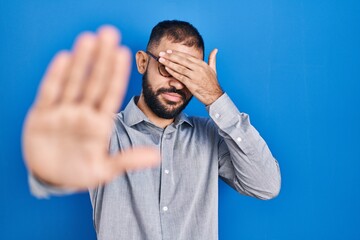 Canvas Print - Middle east man with beard standing over blue background covering eyes with hands and doing stop gesture with sad and fear expression. embarrassed and negative concept.