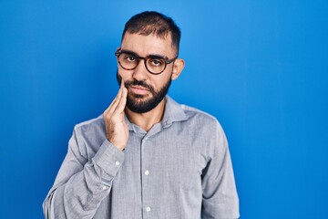 Poster - Middle east man with beard standing over blue background touching mouth with hand with painful expression because of toothache or dental illness on teeth. dentist