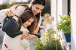 © Aleksandr - Happy daughter watering plants with her beautiful mom. Girl's in bright overall gardening on balcony