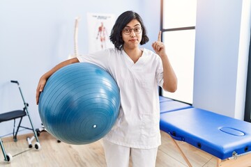 Wall Mural - Young hispanic physiotherapist woman holding pilates ball smiling with an idea or question pointing finger with happy face, number one