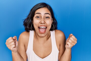 Sticker - Young hispanic woman standing over blue background celebrating surprised and amazed for success with arms raised and open eyes. winner concept.