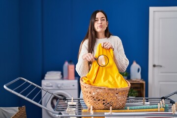 Sticker - Young brunette woman holding magnifying glass looking for stain at clothes puffing cheeks with funny face. mouth inflated with air, catching air.