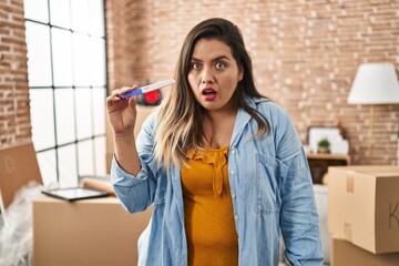 Wall Mural - Young hispanic woman holding pregnancy test result at new home scared and amazed with open mouth for surprise, disbelief face