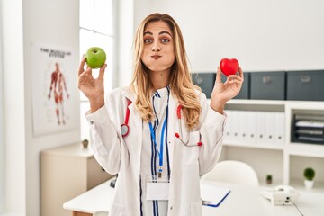 Wall Mural - Young blonde doctor woman holding heart and green apple puffing cheeks with funny face. mouth inflated with air, catching air.