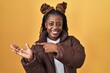 © Krakenimages.com - African woman with braided hair standing over yellow background amazed and smiling to the camera while presenting with hand and pointing with finger.