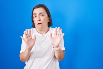 Wall Mural - Middle age hispanic woman standing over blue background moving away hands palms showing refusal and denial with afraid and disgusting expression. stop and forbidden.