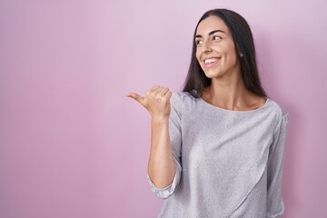 Poster - Young brunette woman standing over pink background smiling with happy face looking and pointing to the side with thumb up.