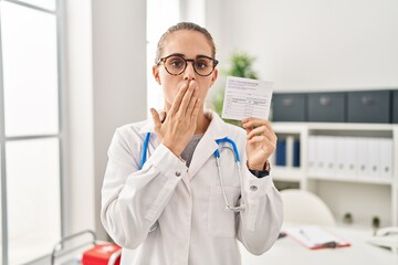 Canvas Print - Young doctor woman holding covid certificate covering mouth with hand, shocked and afraid for mistake. surprised expression