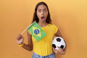 Wall Mural - Young hispanic woman holding brazil flag and football ball in shock face, looking skeptical and sarcastic, surprised with open mouth