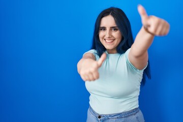 Poster - Young modern girl with blue hair standing over blue background approving doing positive gesture with hand, thumbs up smiling and happy for success. winner gesture.