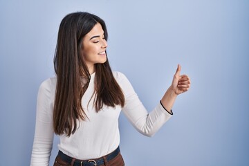 Canvas Print - Young brunette woman standing over blue background looking proud, smiling doing thumbs up gesture to the side