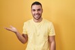 © Krakenimages.com - Young hispanic man standing over yellow background smiling cheerful presenting and pointing with palm of hand looking at the camera.