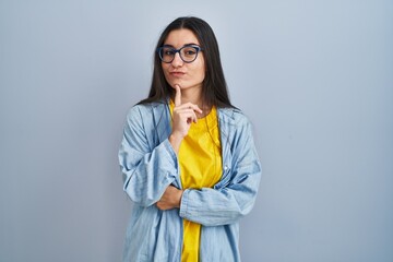 Poster - Young hispanic woman standing over blue background thinking concentrated about doubt with finger on chin and looking up wondering