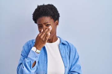 Sticker - African american woman standing over blue background smelling something stinky and disgusting, intolerable smell, holding breath with fingers on nose. bad smell