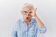 Senior woman with grey hair standing over white background doing ok gesture shocked with surprised face, eye looking through fingers. unbelieving expression.