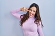 © Krakenimages.com - Young brunette woman standing over blue background doing peace symbol with fingers over face, smiling cheerful showing victory