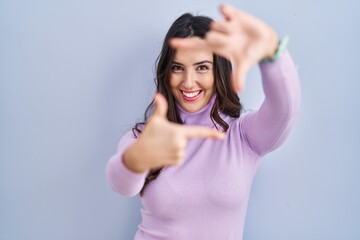 Canvas Print - Young brunette woman standing over blue background smiling making frame with hands and fingers with happy face. creativity and photography concept.