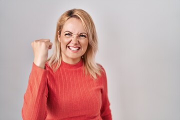 Wall Mural - Blonde woman standing over isolated background angry and mad raising fist frustrated and furious while shouting with anger. rage and aggressive concept.